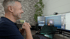 Dr. Marc Dumkow, lachend sitzt vor zwei Monitoren mit einer Videokonferenz und einer Präsentation.