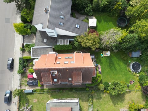Luftaufnahme eines roten Backsteinhauses mit Dachfenstern, umgeben von einem grünen Garten mit Trampolinen und Bäumen.