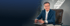 Steuerberater Steffen Rehling in blauem Anzug mit Brille sitzt an einem Schreibtisch vor dunklem Hintergrund.
