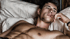 Young man with dark hair and green eyes, bare-chested, lies on a white pillow with a beaded necklace.