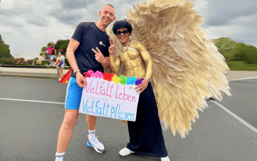 Two people, one with angel wings, hold a sign reading “Live diversity, care for diversity”.