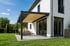 Moderne Terrasse mit Pergola, beige Markise, Gartenmöbeln und Schaukel vor einem weißen Haus.