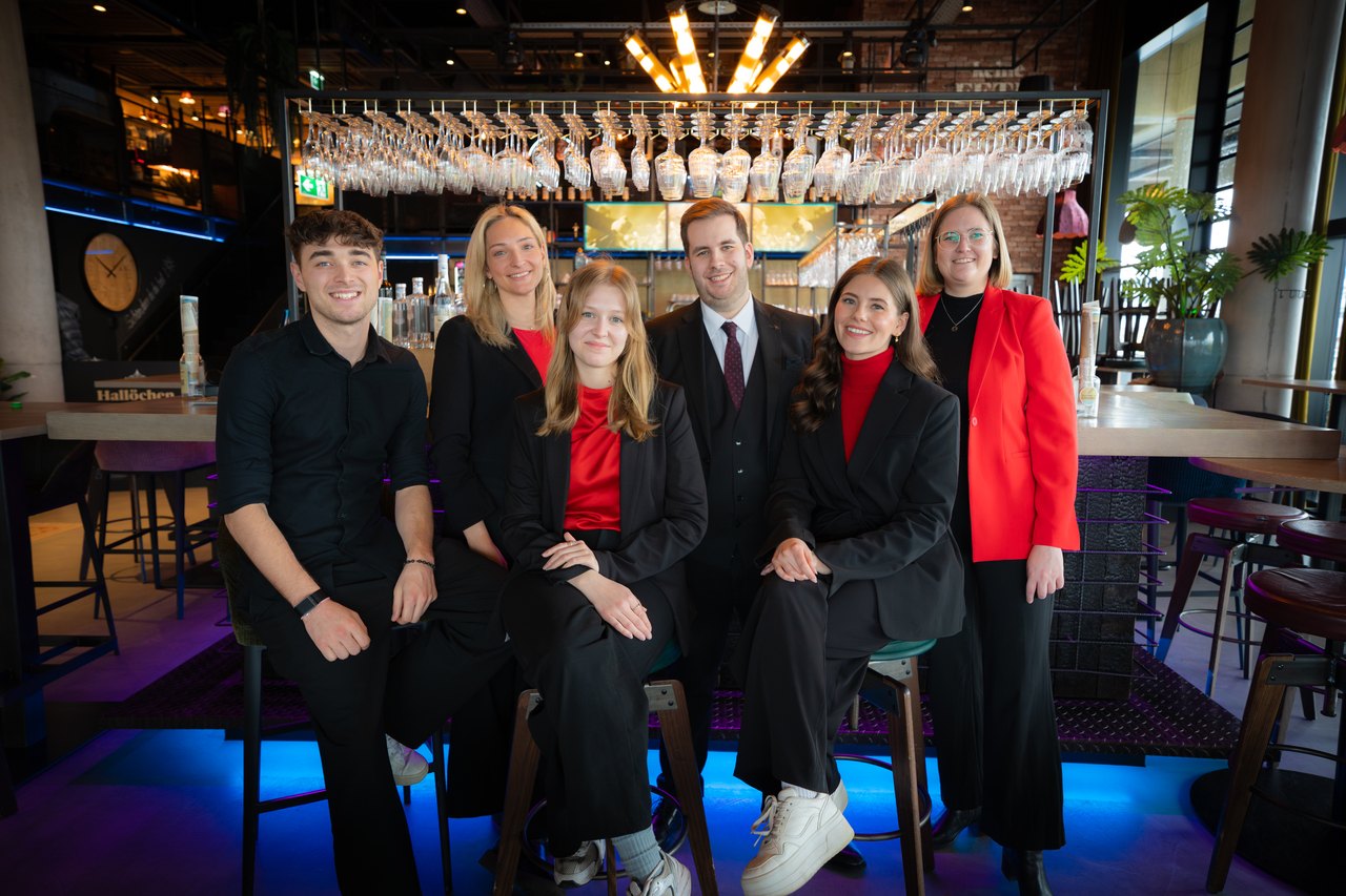 Sechs junge Erwachsene in schwarzer und roter Kleidung lächeln in einer modernen Bar.