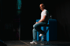 Man in white t-shirt and jeans sits on a high stool on a dark stage with a blue table.