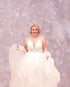 Lächelnde Frau in einem weißen Hochzeitskleid mit Spitzenoberteil vor einem glitzernden, hellrosa Bokeh-Hintergrund.