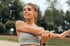 A smiling, athletic young woman stretching her arm outdoors.