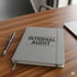 Graues Notizbuch mit der Aufschrift „INTERNAL AUDIT“ liegt neben einem schwarzen Stift auf einem Holzschreibtisch.
