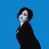 Webdesign Nürnberg TN Production -Leticia Neumann in einem schwarzen Blazer mit dunklen Haaren und Make-up blickt in die Kamera vor blauem Hintergrund.