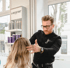 Ein Friseur mit Brille stylt die Haare einer blonden Kundin vor einem Regal mit La Biosthétique Produkten.