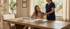 A nurse in blue scrubs shows a tablet to a seated woman at a wooden table in a home setting.