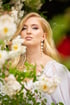 Blonde Frau mit Perlenohrringen und weißem Kleid posiert hinter blühenden weißen Rosen im Freien.