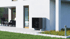 Eine schwarze Wärmepumpe an der weißen Außenwand eines modernen Hauses, mit Terrasse und Garten.