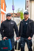Zwei Männer in schwarzen MMS-Jacken und Mützen, halten Werkzeugkoffer, stehen vor Häusern.