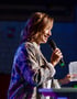 A woman in a patterned jacket and velvet pants speaks into a microphone on stage, holding papers.