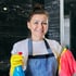 Lächelnde Reinigungskraft mit Schürze, gelben Handschuhen, Sprühflasche und Putztuch vor einer schwarzen Marmorwand.