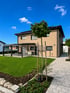 Modernes Einfamilienhaus mit beiger Fassade, grüner Rasenfläche, gepflastertem Weg und einem jungen Baum unter blauem Himmel.