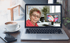 Eine Frau mit Brille und rosa Hoodie lächelt von einem Laptop-Bildschirm; daneben eine Tasse Kaffee und ein Smartphone.