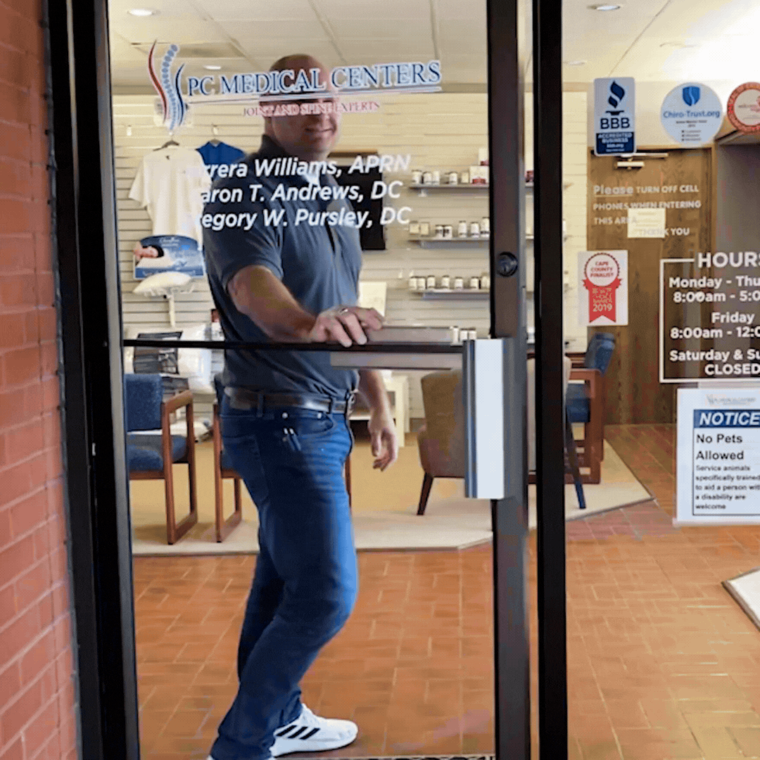 PC Medical Centers staff member opening the clinic's glass front door, with the practice logo and provider names on the glass and the waiting area behind