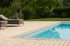 Moderner Außenpool mit hellblauem Wasser, umrahmt von einer hellen Holzterrasse, Sonnenliegen und grüner Gartenlandschaft.