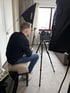 Ein Mann sitzt auf einem Hocker und blickt auf eine Videokamera auf einem Stativ, umgeben von zwei Softboxen.