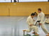 Zwei Männer in weißen Karate-Anzügen trainieren in einer Turnhalle; einer kniet, der andere steht.
