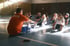Trainer im orangefarbenen Shirt macht mit einer Gruppe lachender Kinder Dehnübungen in einer Turnhalle.