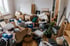 Unordentliches Zimmer mit zahlreichen Umzugskartons, Müllsäcken, einer Matratze und verstreuten Haushaltsgegenständen.