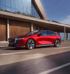 Roter Mazda EZ-6 Elektroauto fährt auf einer Straße vor einem modernen Gebäude.