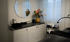 Moderner Behandlungsraum mit weißem Sideboard, schwarzem Waschbecken, rundem Spiegel, bunten Blumen und Kosmetikprodukten.