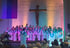 Ein Chor in weißen und blauen Roben singt in einer Kirche unter einem Kruzifix und einem Regenbogenbanner.