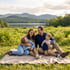 Smiling family of four sits on a blanket in a green field with a lake and mountains in the background.