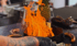 Koch mit Handschuhen grillt Würstchen auf flacher Grillplatte mit Holzfeuer und Ananas im Hintergrund.