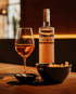 Ein Glas Roséwein, eine Flasche "bress" und zwei schwarze Schalen mit Snacks auf einem Holztisch.