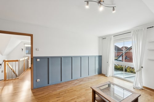 Helles Wohnzimmer mit Holzfußboden, blauer Wandverkleidung, Fenster mit weißem Vorhang und Glastisch.