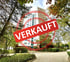 Hohes Wohngebäude umgeben von Bäumen und Sträuchern, mit rotem Stempel "VERKAUFT" im Vordergrund.