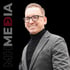 Lachender Mann mit Brille, schwarzem Rollkragenpullover und grauem Sakko vor schwarzem Hintergrund mit "MHMEDIA" Text.
