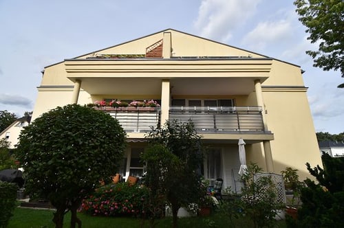 Gelbes Wohnhaus mit zwei Balkonen und Blumenkästen, umgeben von Bäumen und Sträuchern unter bewölktem Himmel.