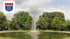 Springbrunnen in einem Parkbecken vor grünen Bäumen mit dem Wappen von Ladbergen oben links.