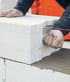 A construction worker places a large white aerated concrete block onto a layer of mortar using a red leveling line.