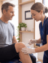 A physical therapist in blue scrubs uses a handheld TENS unit on a patient lying on a treatment table.