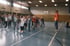 Ein Trainer in orangefarbenem Shirt leitet eine Gruppe Kinder beim Sportunterricht in einer Turnhalle an.