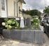 Ein modernes Hochbeet aus grauen Beton-L-Steinen mit grünen Hortensien und einer Wetterfahne vor einem Wohnhaus.