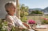 Seniorin sitzt lächelnd in der Sonne auf einem Balkon neben einer Medikamentenbox und einem Glas Wasser.
