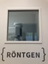 Weiße Wand mit einem Fenster, durch das ein Röntgengerät sichtbar ist, und dem Wort "RÖNTGEN" darunter.