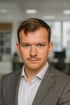 Lukas Förster mit braunem Haar und Schnurrbart trägt einen grauen Anzug und ein weißes Hemd.