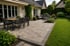 Gepflasterte Terrasse mit Esstisch, Lounge-Möbeln und angrenzendem grünen Rasen in einem gepflegten Garten.