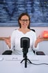 Eine lächelnde Frau mit Brille sitzt vor einem Mikrofon und gestikuliert mit den Händen.