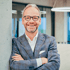 Frank Möller, professionelles Business-Porträt eines lächelnden Mannes im blauen Anzug vor einem modernen Bürohintergrund.