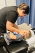 A chiropractor in black scrubs performs a neck adjustment on a young boy lying on a clinical table holding a blue toy.