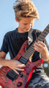 A boy with blonde hair plays a red electric guitar with black pickups.